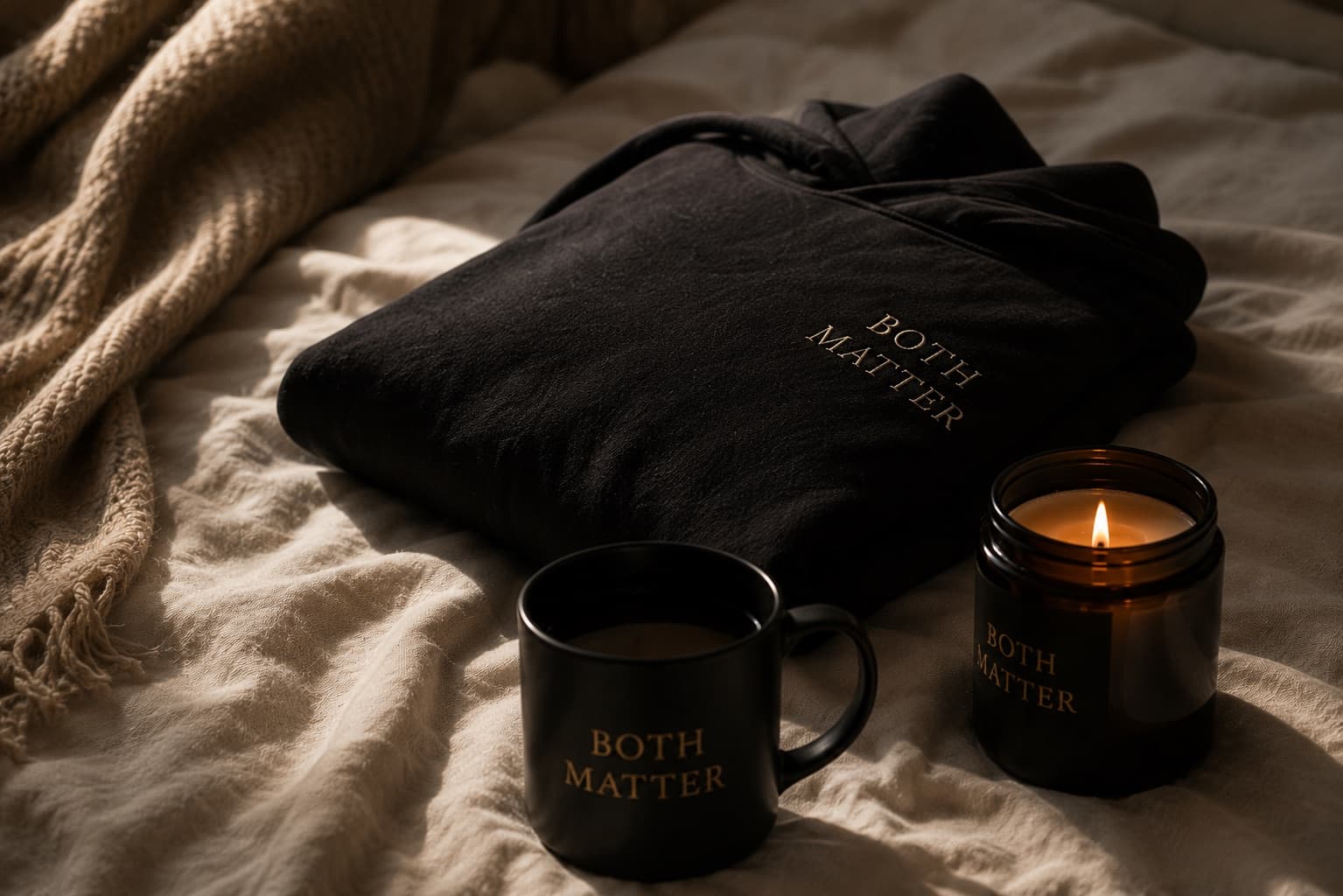 Folded Both Matter hoodie with a mug and candle on soft bedding.